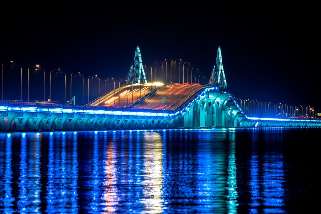 Pensacola Bay Bridge illuminated in blue at night, with light trails and colorful reflections across the water
